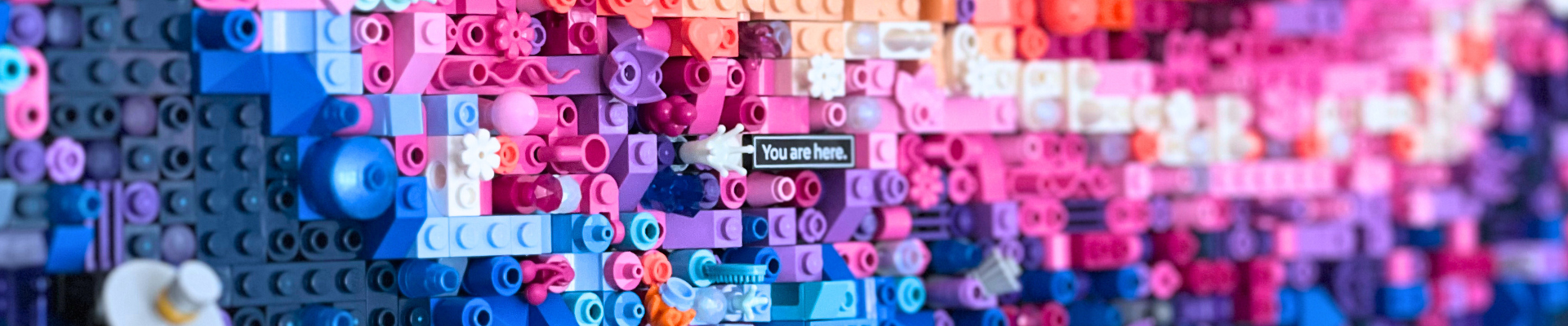 Ausschnitt aus dem Lego-Motiv der Milchstraße. Viele unterschiedliche Lego-Steine in den Farben blau, pink, weiß und orange wechseln sich ab. In der Bildmitte steht „You are here.“ auf einem Stein.